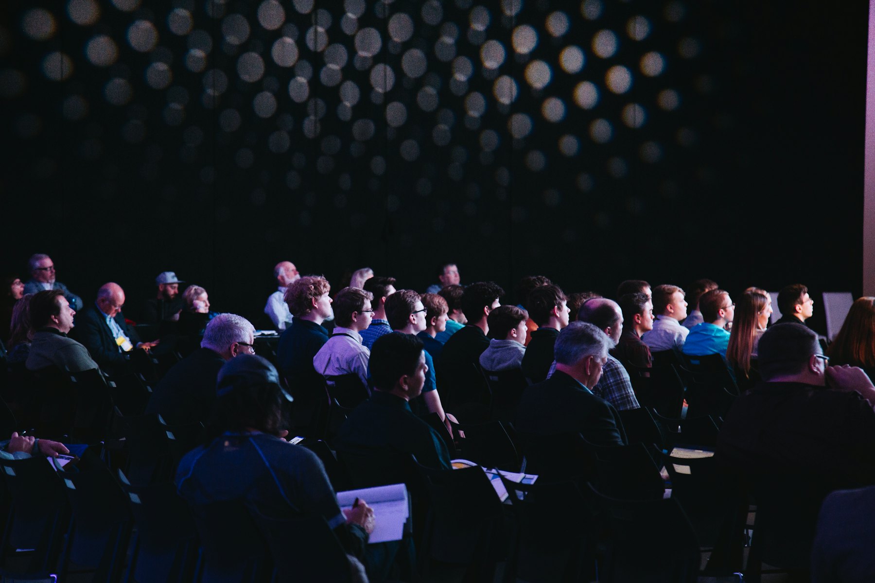 A seated audience in a darkened conference hall, waiting for a session to begin